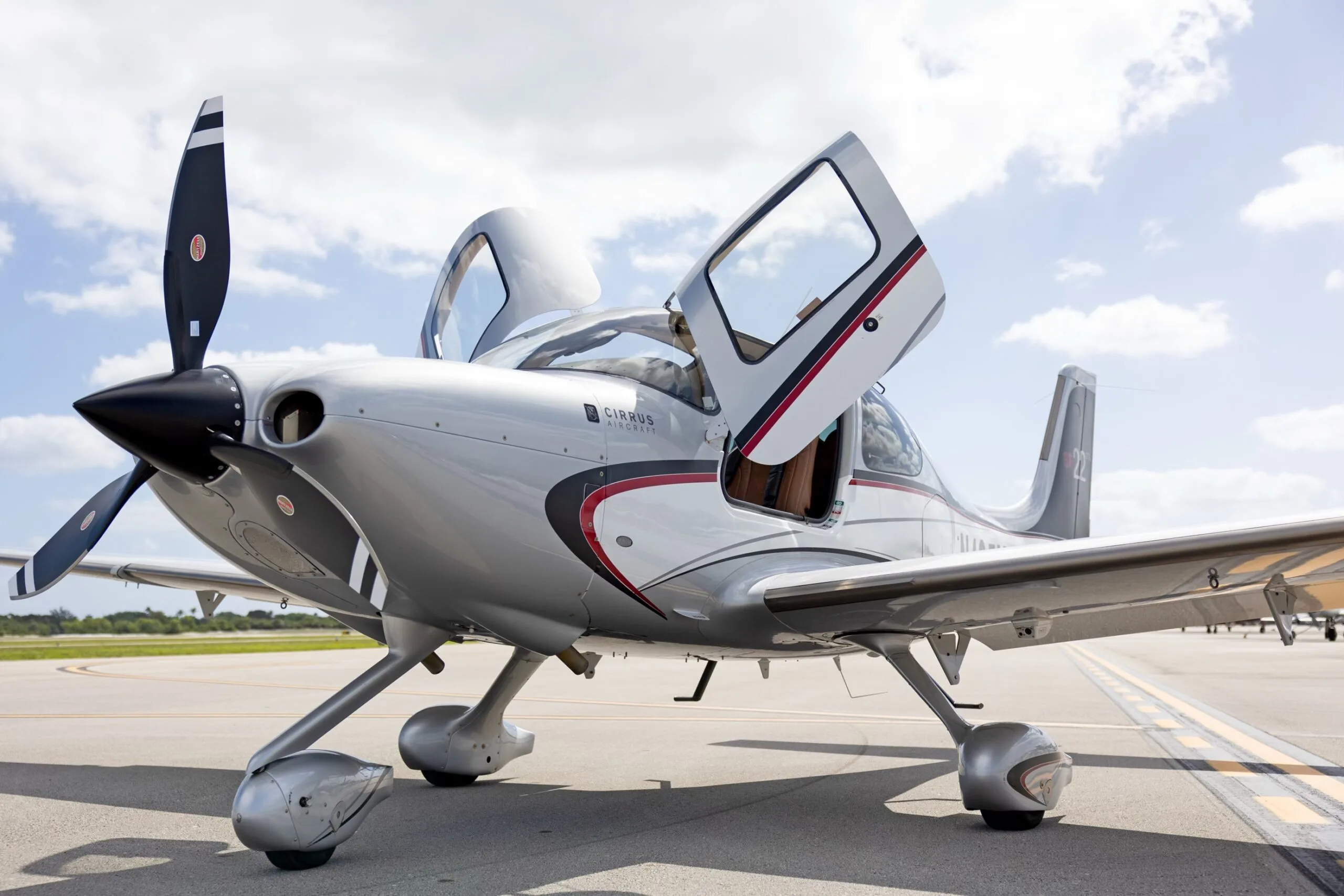 A silver Cirrus SR22 GTS aircraft with red and black accents, doors open, parked on an airfield under a partly cloudy sky.