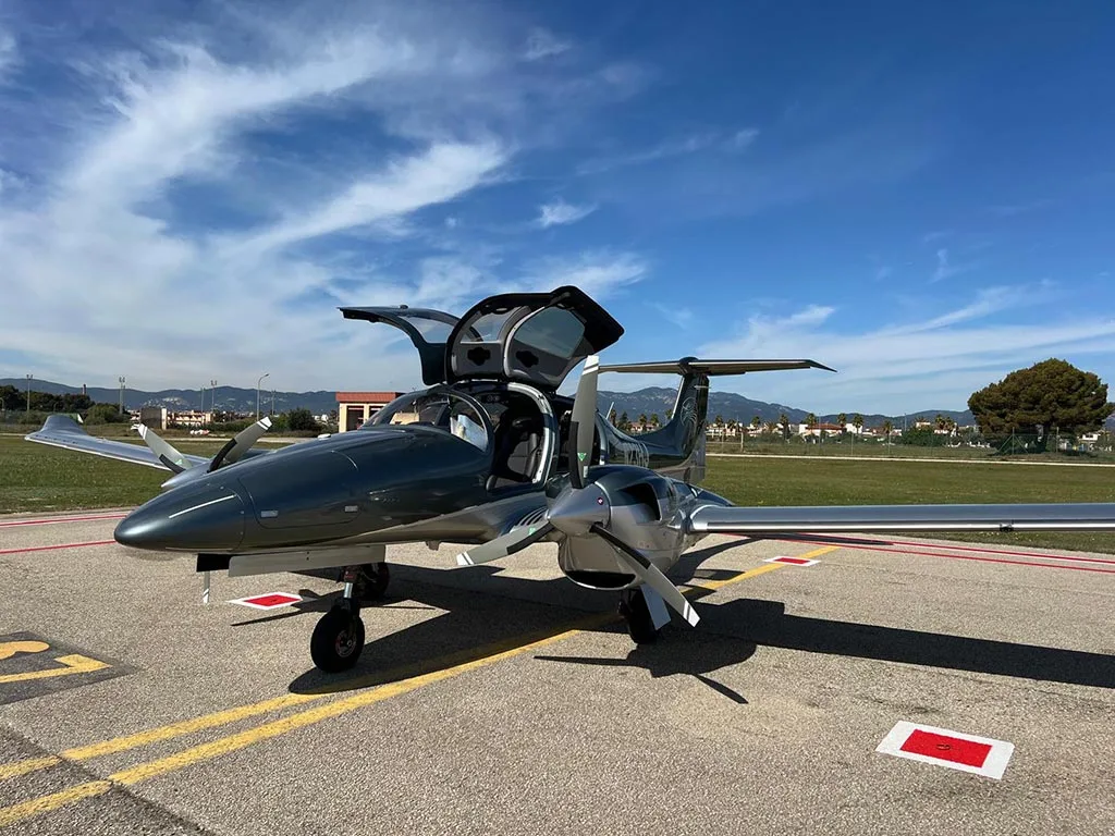 A front-quarter view of a dark grey Diamond DA62 aircraft with its gull-wing doors open, parked on an airport tarmac under a blue sky.
