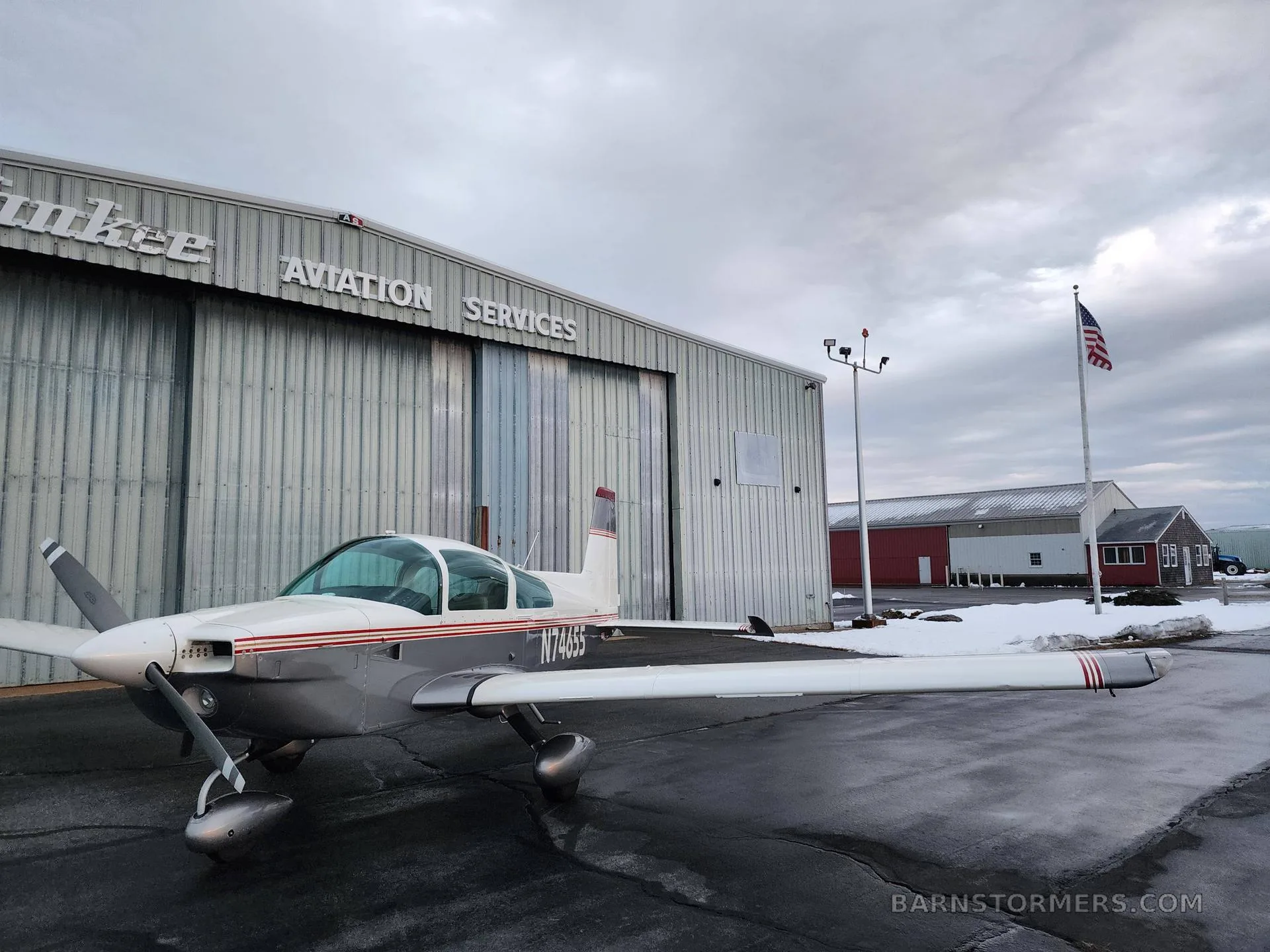 A white and silver Grumman AA-5 Traveler aircraft with red stripes parked on a wet tarmac, with an American flag in the background.