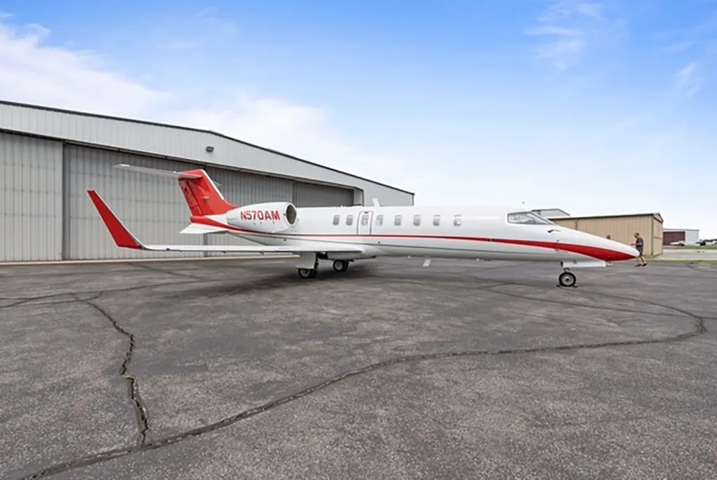 A white and red business jet (N570AM) parked on an asphalt tarmac next to a hangar.