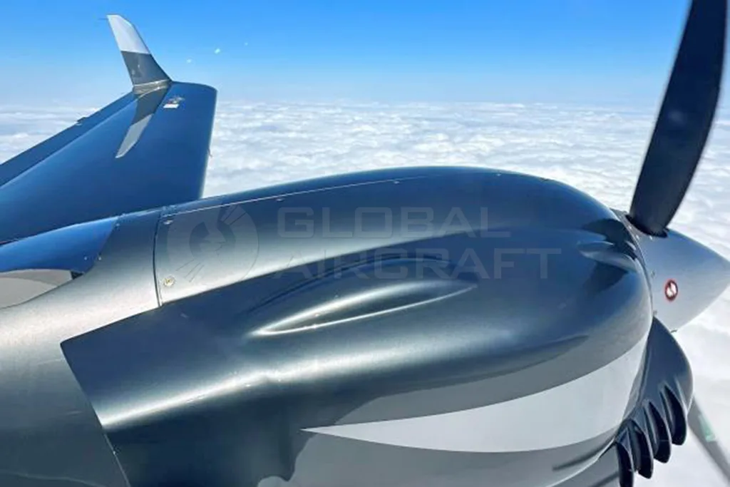 A close-up view of the dark grey engine nacelle and propeller of a Diamond DA62 aircraft flying above a layer of white clouds.