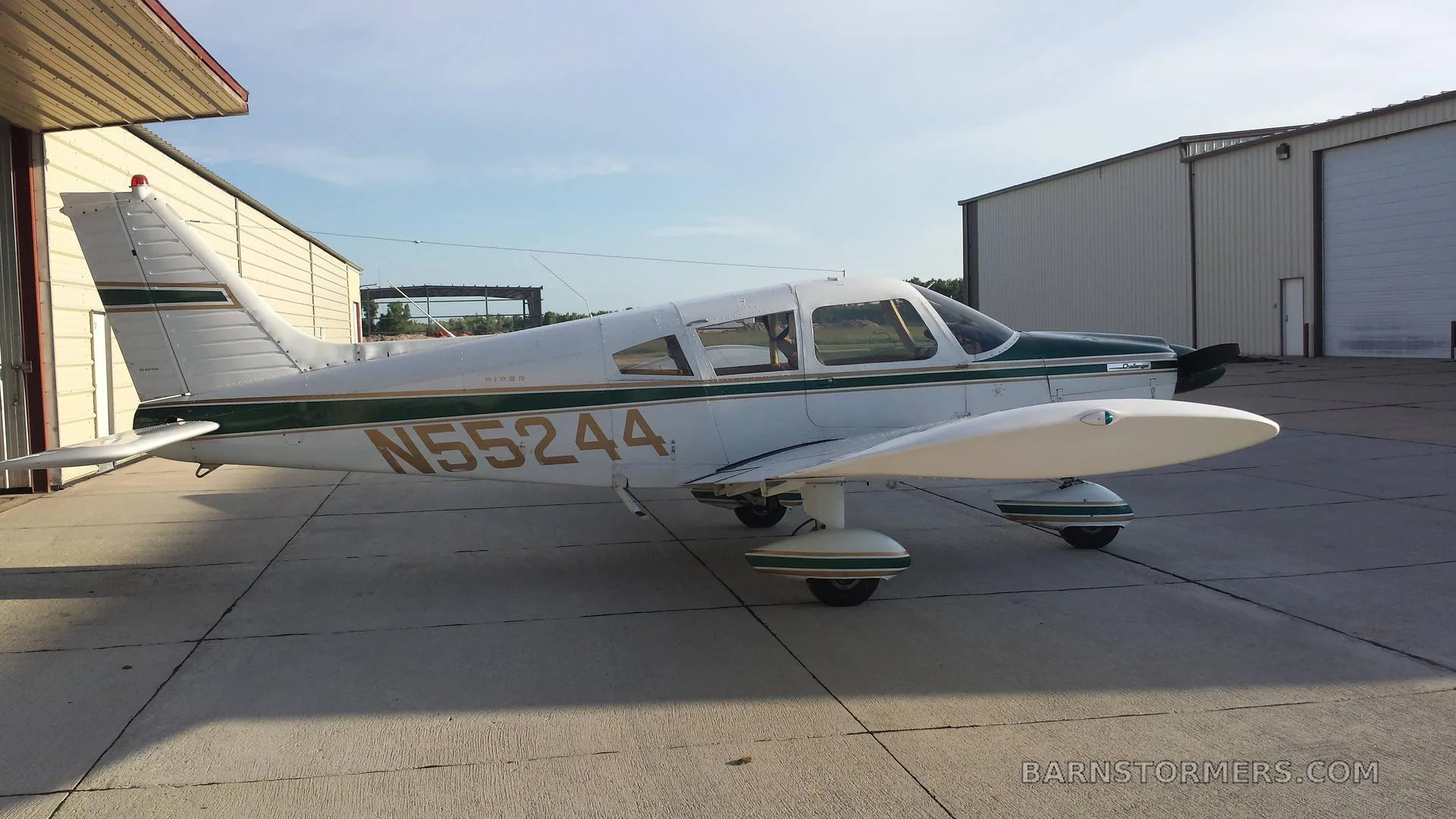 A white and green Piper Cherokee Challenger (N55244) parked on a concrete ramp next to hangars.