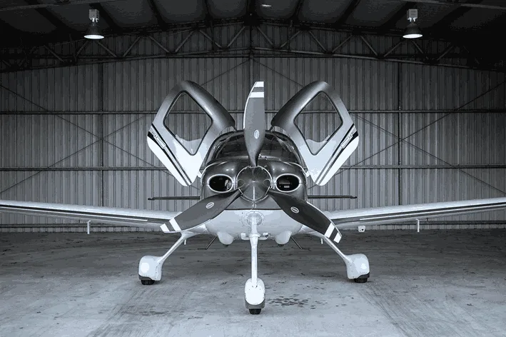 A light-colored propeller aircraft with open gull-wing doors is parked head-on inside a hangar.