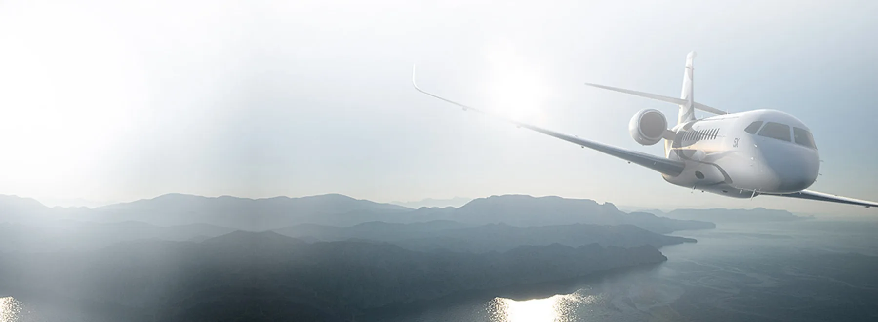 A generic white business jet flying over a misty mountainous coastline with the sun glinting off its wing.