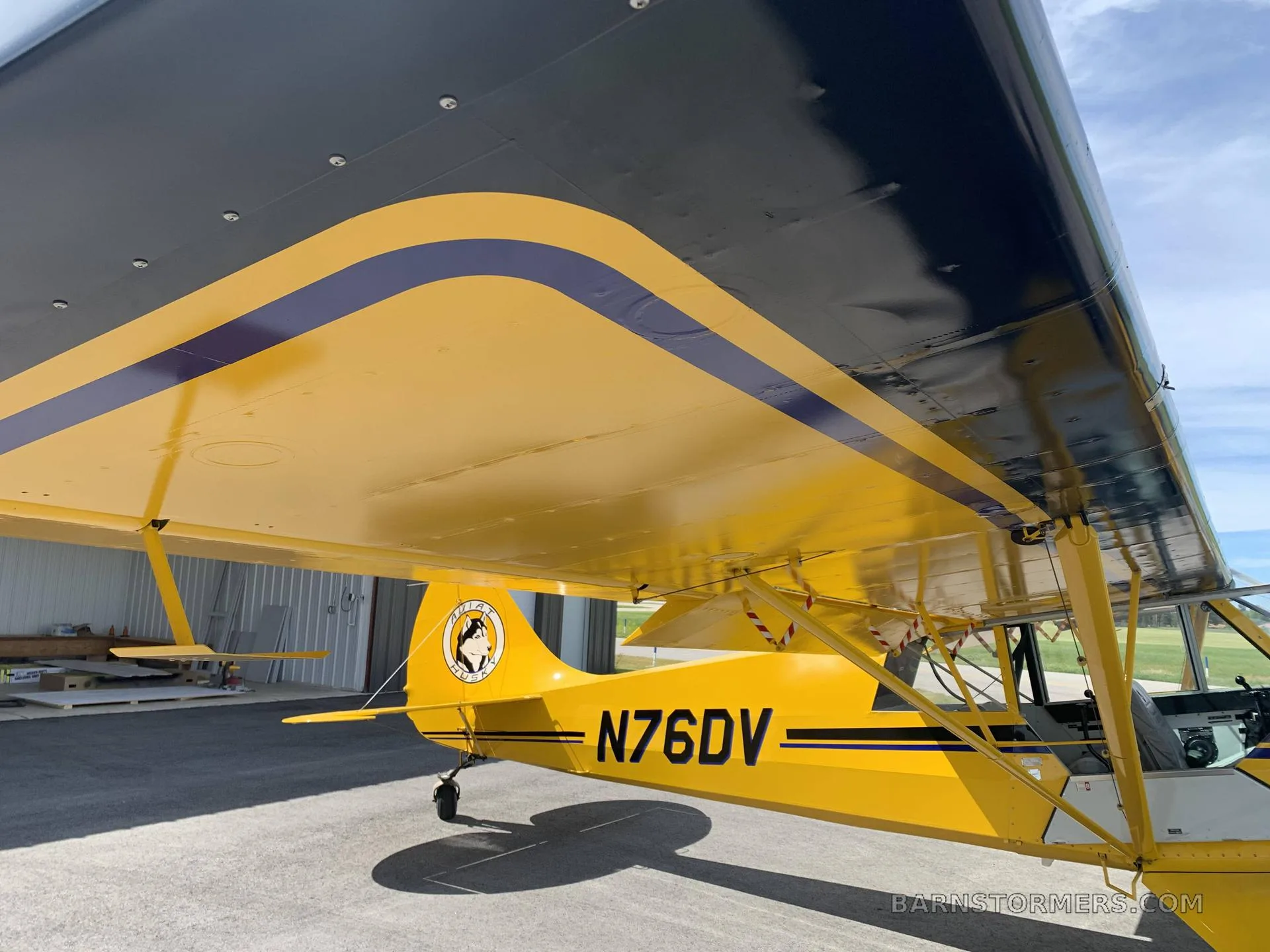 A close-up view of the yellow and black wing and tail of an Aviat Husky A-1 aircraft with registration N76DV, parked in a hangar.