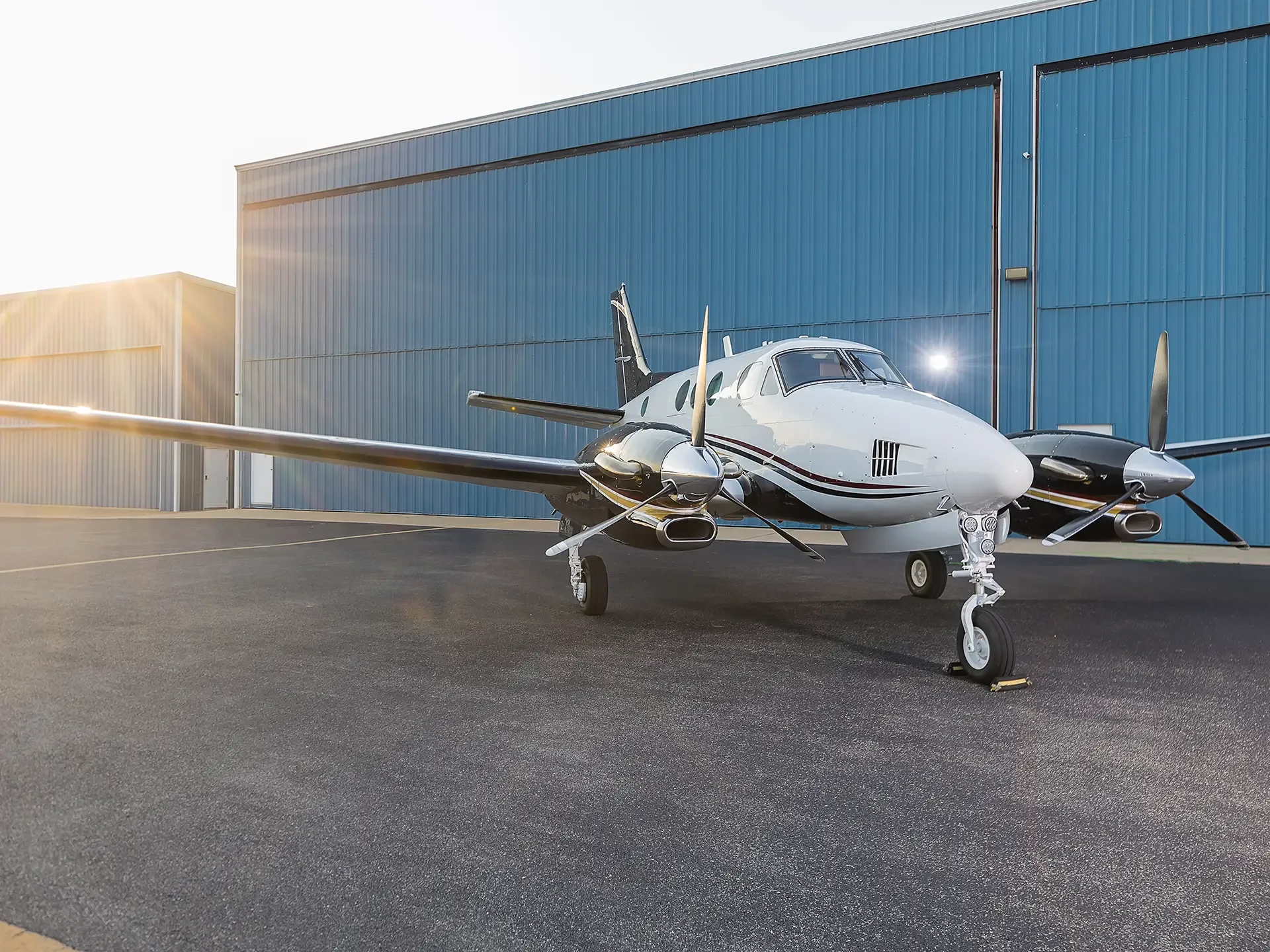 A white and black twin-engine turboprop aircraft with red stripes is parked on an asphalt ramp in front of blue hangars at sunset.