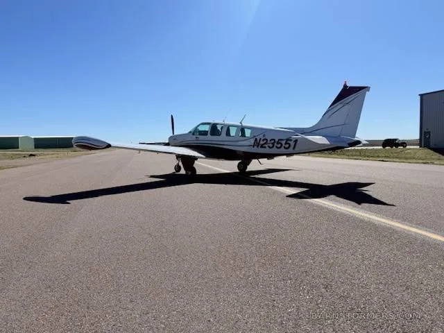 A white and red Beechcraft Bonanza aircraft is parked on an asphalt runway, viewed from the rear-left.