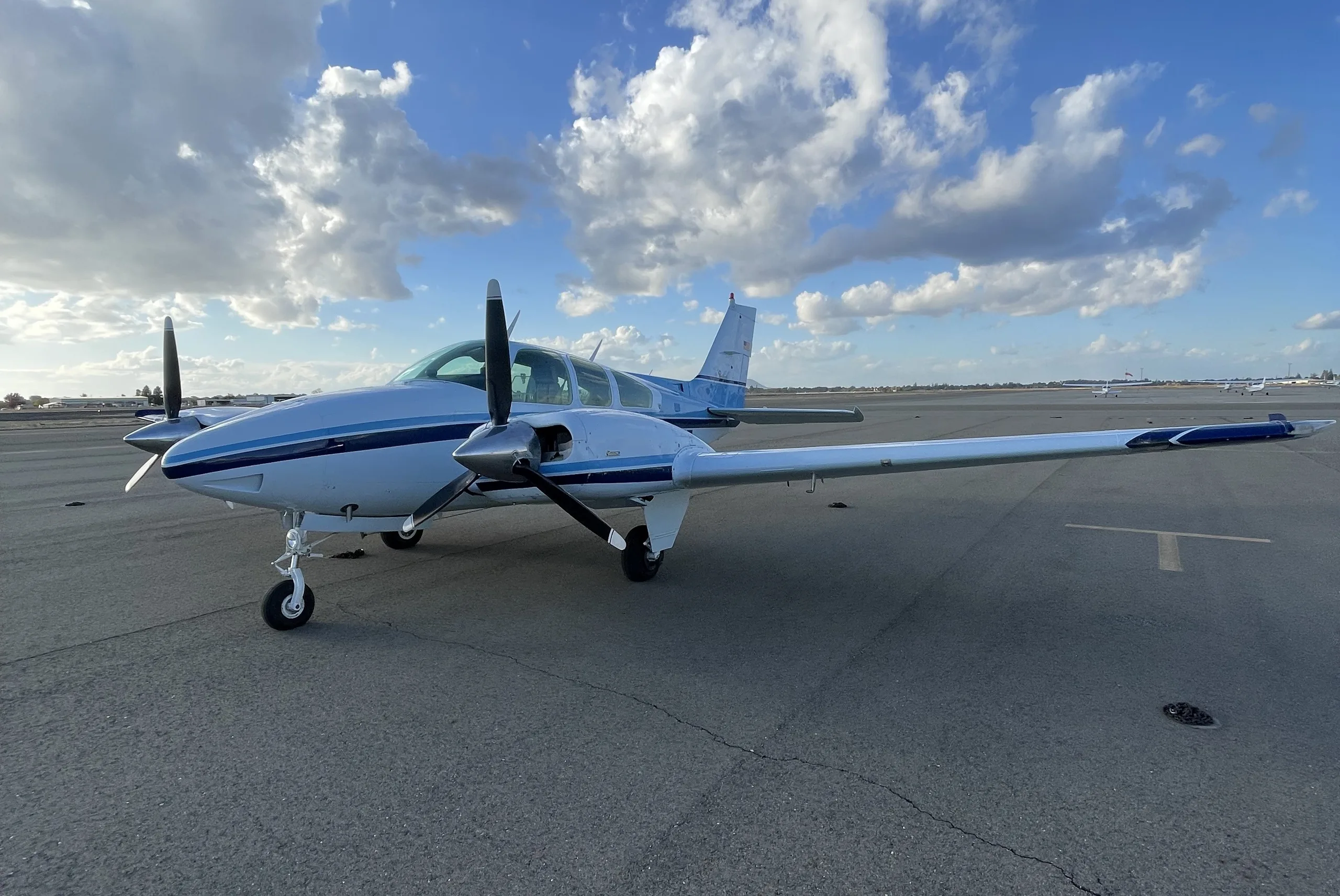 A white and blue twin-engine propeller aircraft is parked on an asphalt tarmac under a partly cloudy sky.