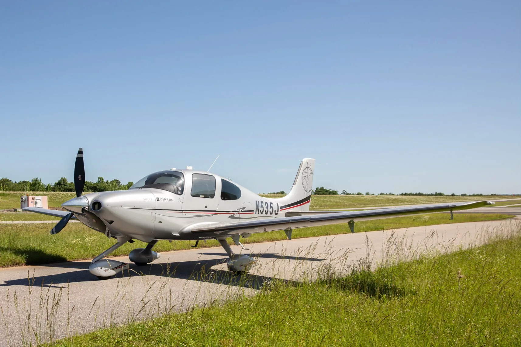 A silver Cirrus SR22T aircraft with registration N535J is parked on an asphalt strip next to green grass under a clear blue sky.