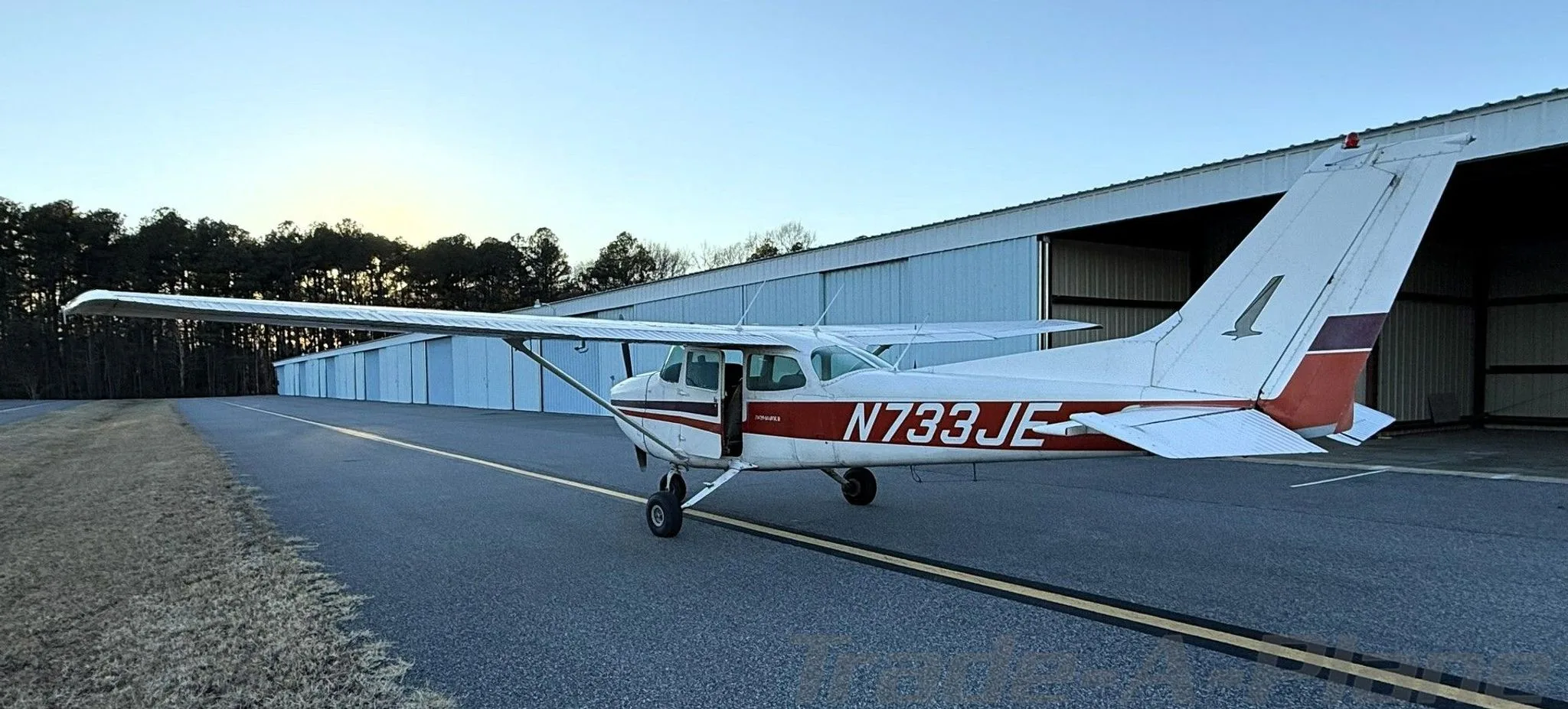 A white Cessna 172N with red and black stripes, registration N733JE, parked on an airfield with a row of hangars behind it.