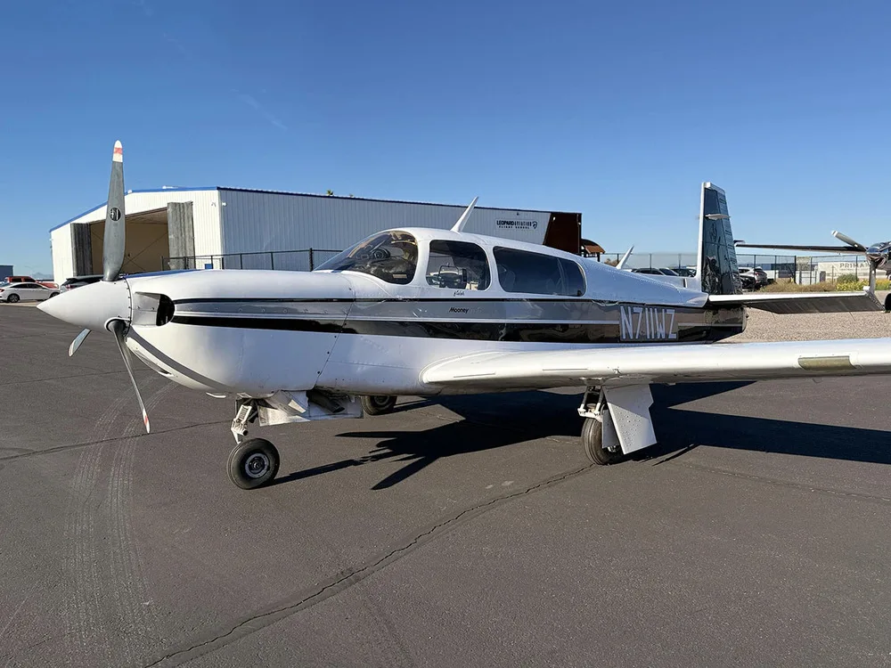 A front-left view of a white Mooney aircraft with black and grey stripes, tail number N711WZ, parked on an airfield.