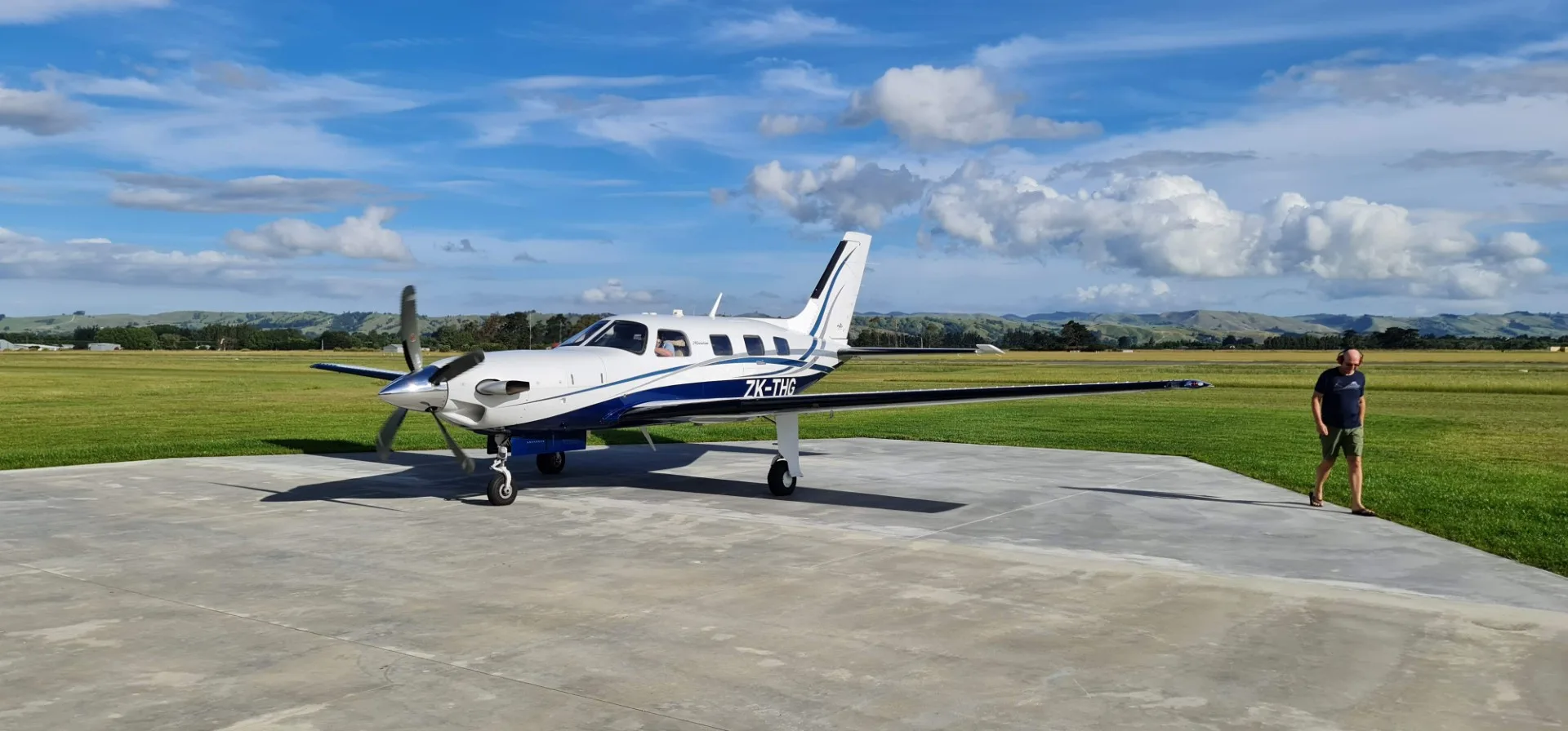 A white and blue Piper M600 aircraft, registration ZK-THG, parked on a concrete pad at an airfield under a partly cloudy sky.