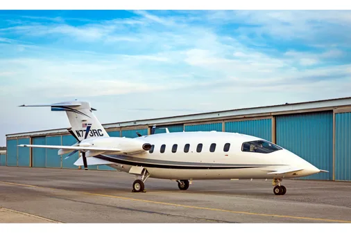 A white Piaggio P.180 Avanti aircraft with blue and grey stripes on the tail, parked on an asphalt tarmac in front of blue hangars under a blue sky.