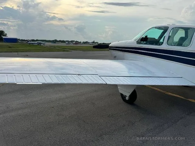 A side view of the wing and front fuselage of a white and blue Beechcraft Bonanza on an airfield, with other aircraft in the distance.