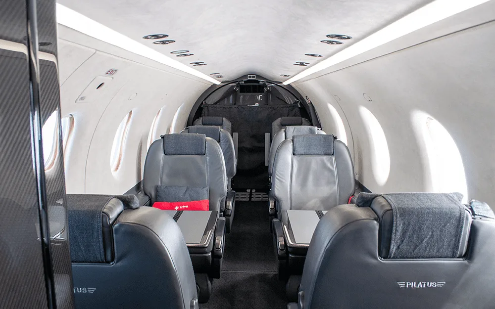 The spacious cabin interior of a Pilatus PC-12 aircraft, showing rows of dark grey leather seats with fold-out tables.
