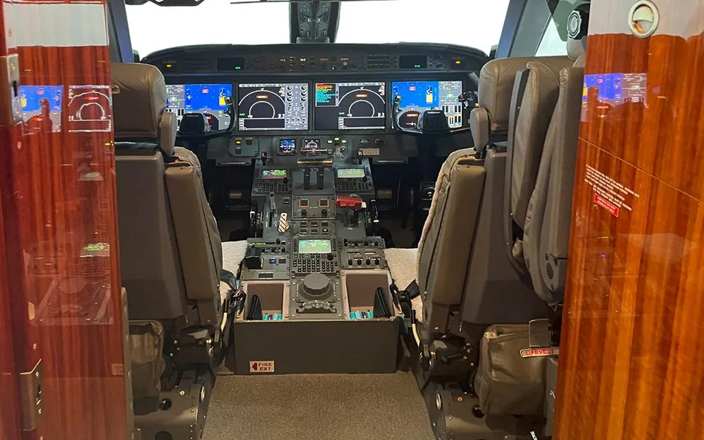 View into the cockpit of a Gulfstream G550, showing the instrument panel with multiple displays and the center console, framed by two pilot seats and wood paneling.
