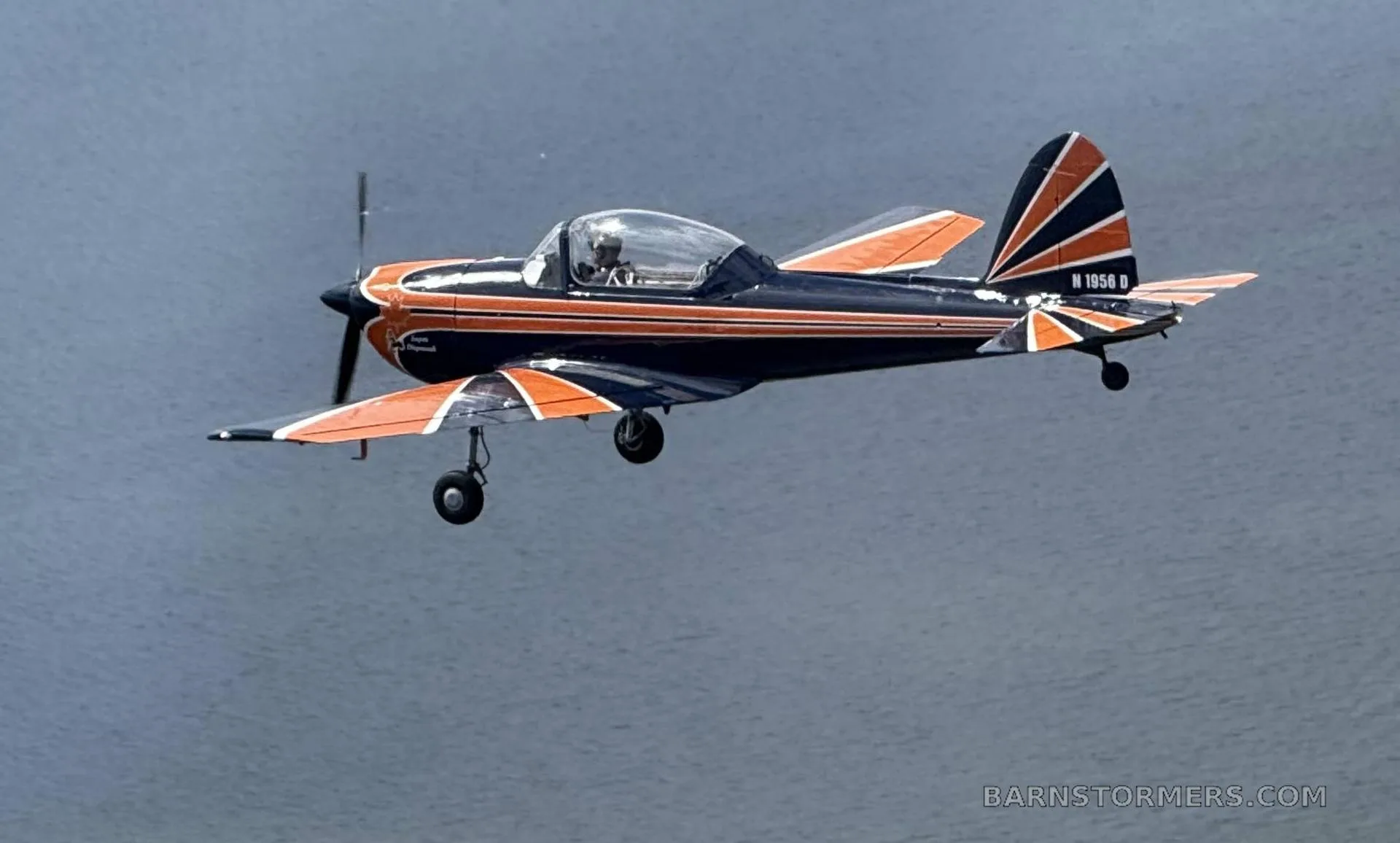 A blue and orange single-engine aircraft with tail number N 1956 D flying low over a body of water.