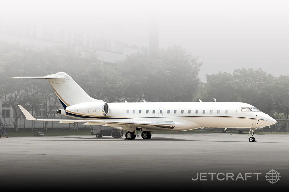 A white business jet with a dark blue and gold stripe on the tail and fuselage is parked on an asphalt tarmac with hazy buildings and trees in the background.