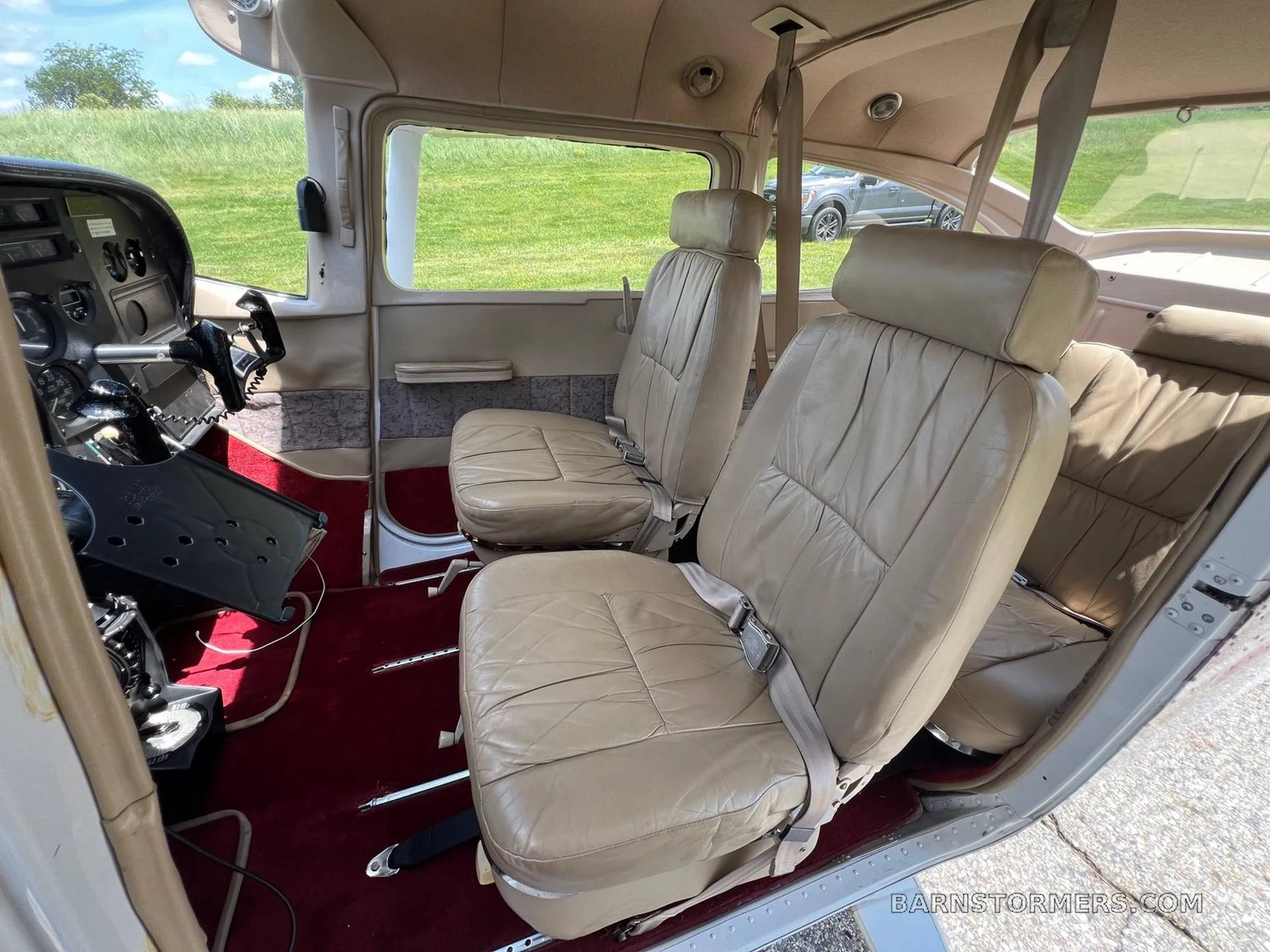 The tan leather interior of a small aircraft cabin with two front seats and a view of the grassy airfield outside.