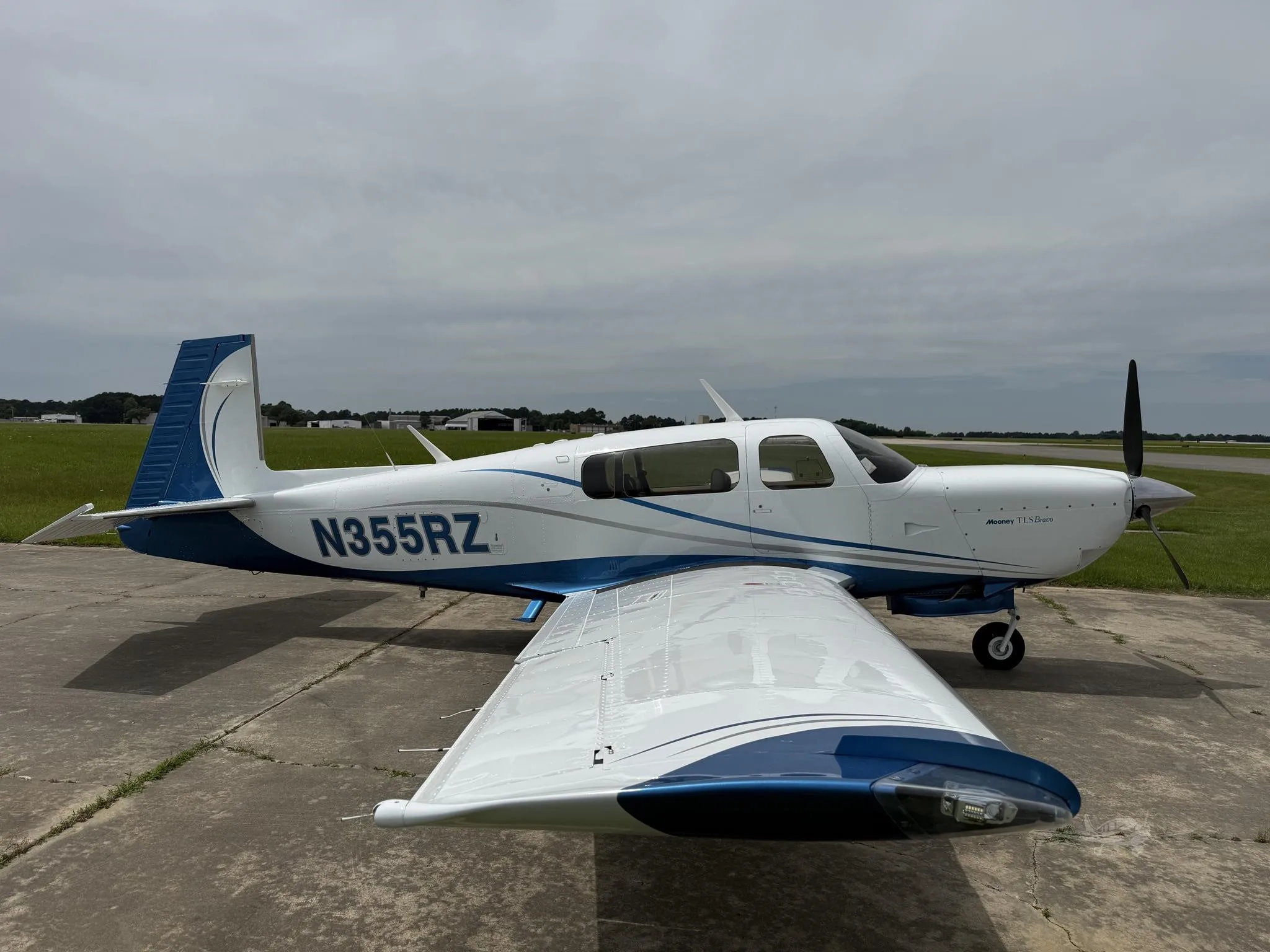 A white Mooney TLS Bravo aircraft with blue and grey accents is parked on a concrete tarmac under a cloudy sky.