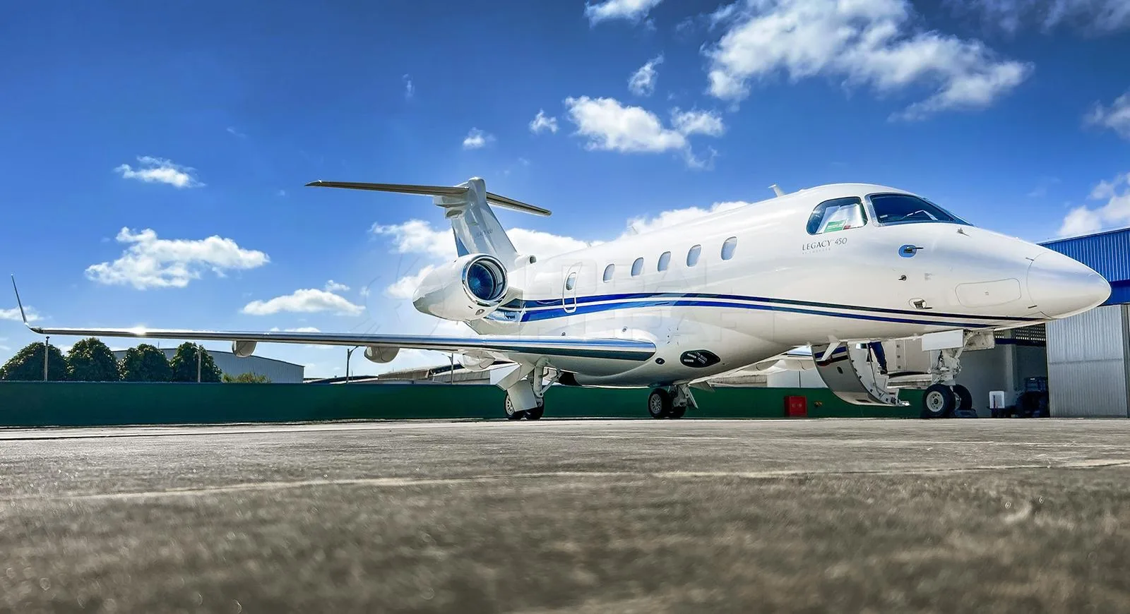 A white Embraer Legacy 450 business jet with blue stripes parked on an asphalt apron under a blue sky.