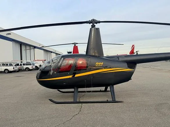 A black Robinson R44 Raven II helicopter with a yellow stripe, parked on an airfield next to a hangar.