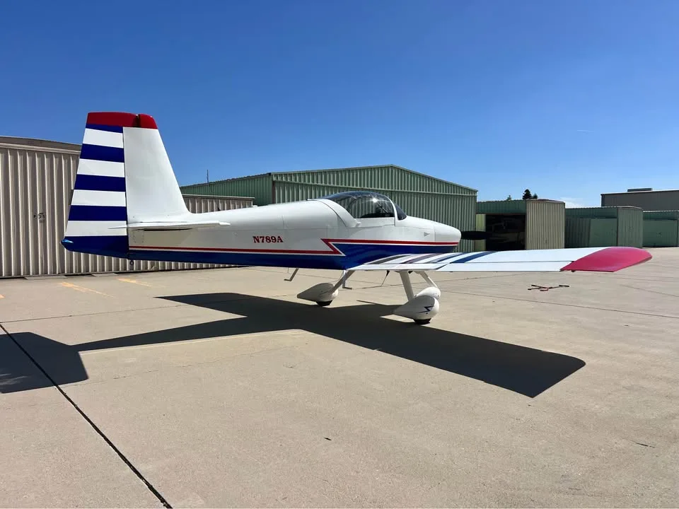 A white, blue, and red single-engine aircraft with a striped tail and wingtips, parked on an airfield under a clear blue sky.