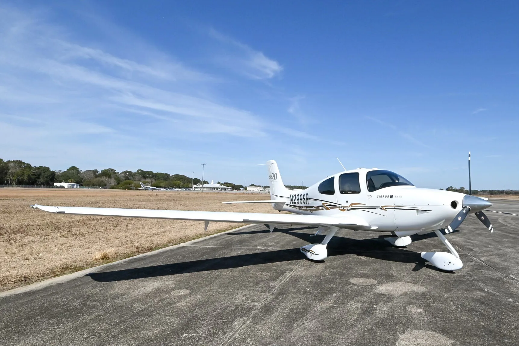 A white Cirrus SR20 aircraft with black and gold accent stripes, registration N299SR, parked on an airfield with buildings in the distance.