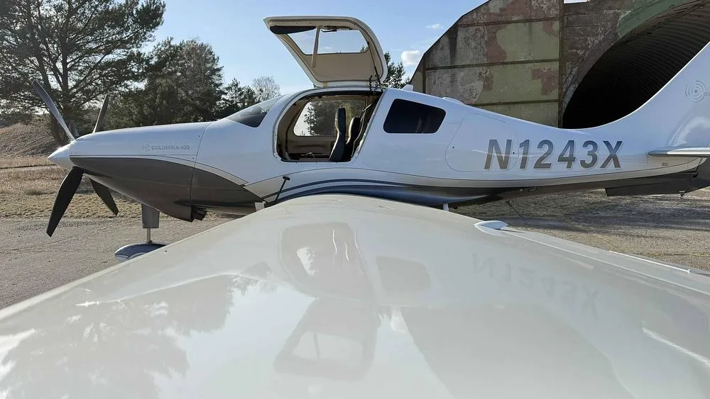 A side view of the white and grey Columbia 400 aircraft N1243X with its doors open, parked near a hangar.