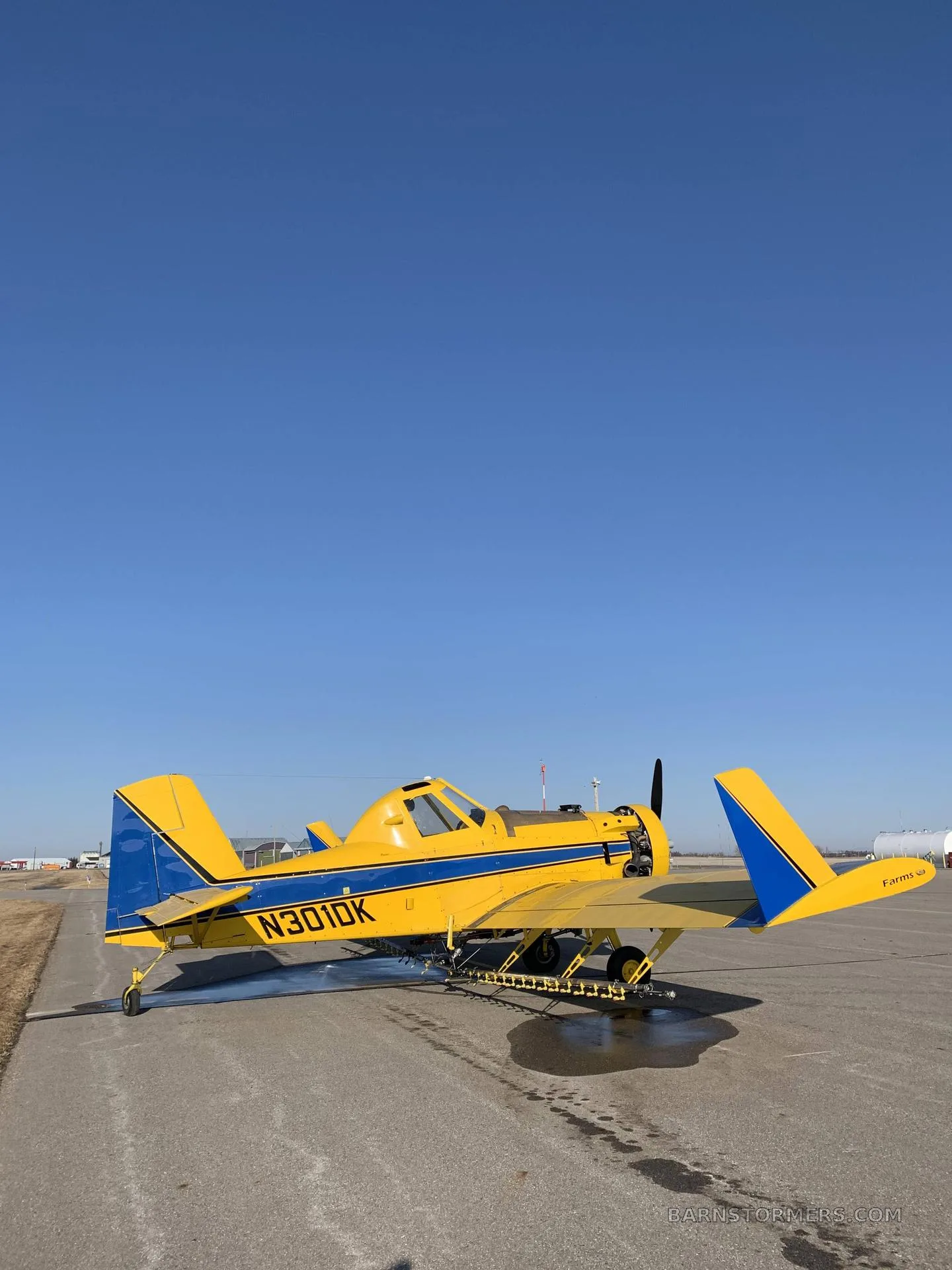A yellow agricultural aircraft with blue stripes and registration N301DK parked on an airfield under a clear blue sky.