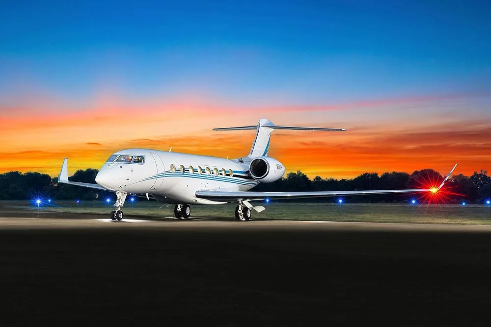 A white Gulfstream G650 business jet with blue stripes on a runway at sunset with landing lights on.