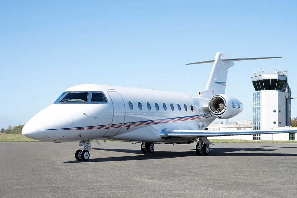 A white business jet, likely a Gulfstream, with blue and red stripes, parked on an asphalt tarmac with a control tower in the background.