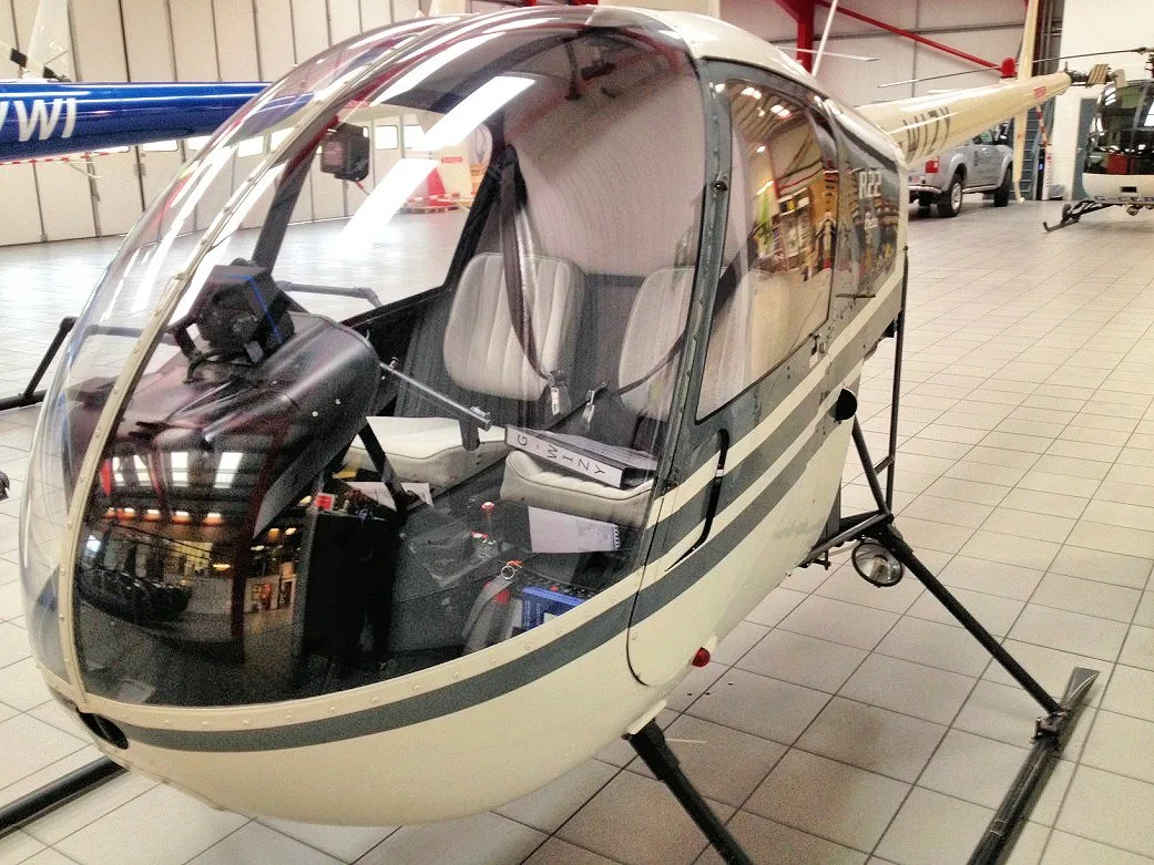 A white helicopter with a dark stripe, possibly a Robinson R22, parked inside a hangar with other aircraft in the background.
