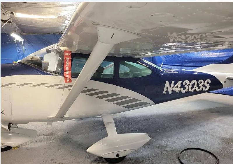 A side view of the blue and white Cessna N4303S's fuselage and wing, showing the tail number and a "REMOVE BEFORE FLIGHT" tag.