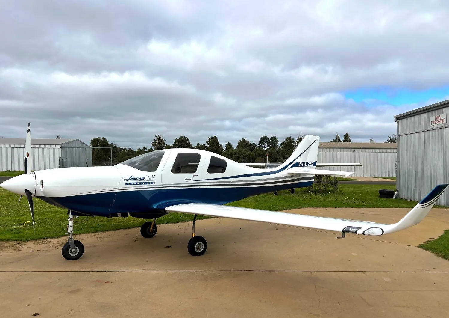 A white and blue Jabiru J430 aircraft (VH-LZS) with a propeller, parked on an asphalt surface next to a hangar under a cloudy sky.