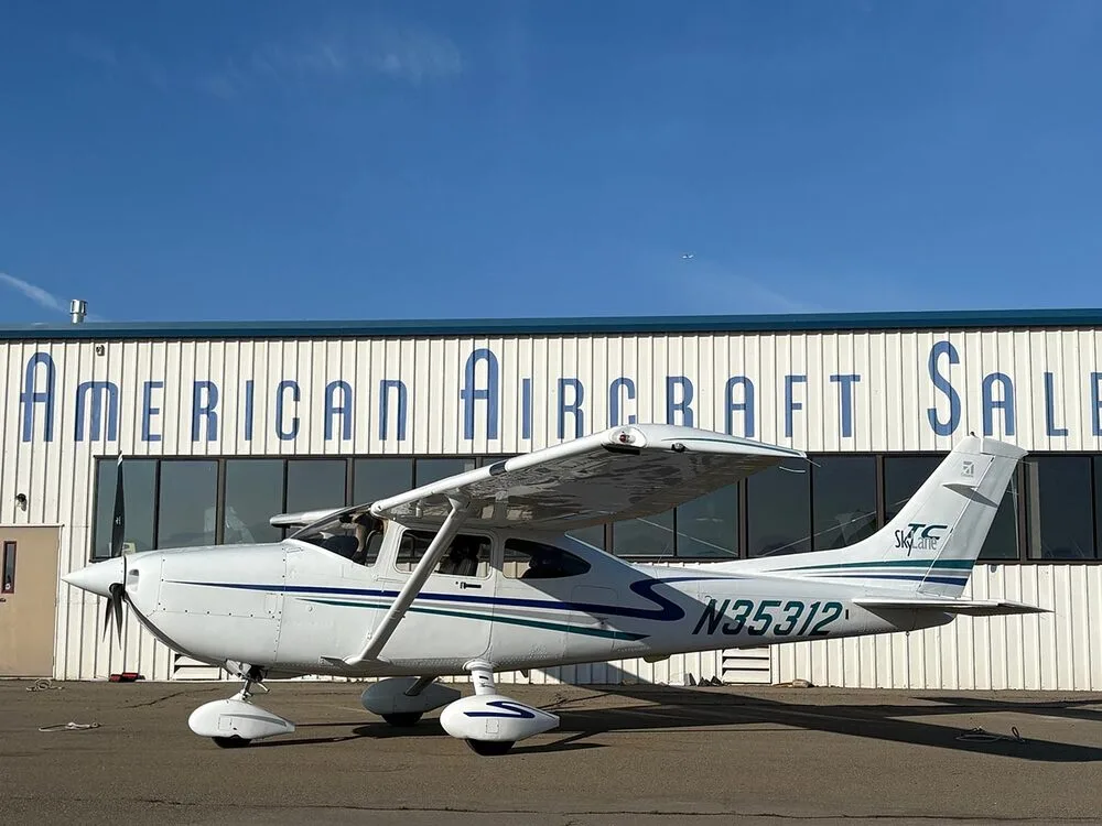 A white Cessna 182 Skylane with blue and green stripes, registration N35312, parked in front of a building labeled "American Aircraft Sale".
