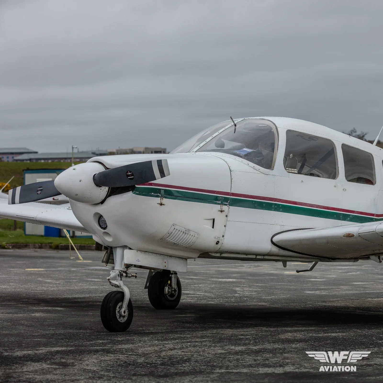 A white single-engine propeller aircraft with green and red stripes on the fuselage, parked on an asphalt tarmac under a cloudy sky.