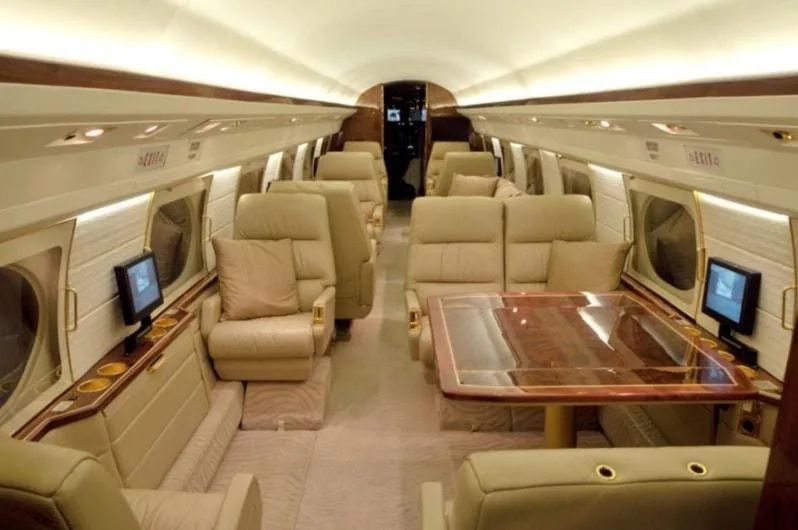 The luxurious interior of a private jet cabin with beige leather seats, a large wooden table, and individual monitors.