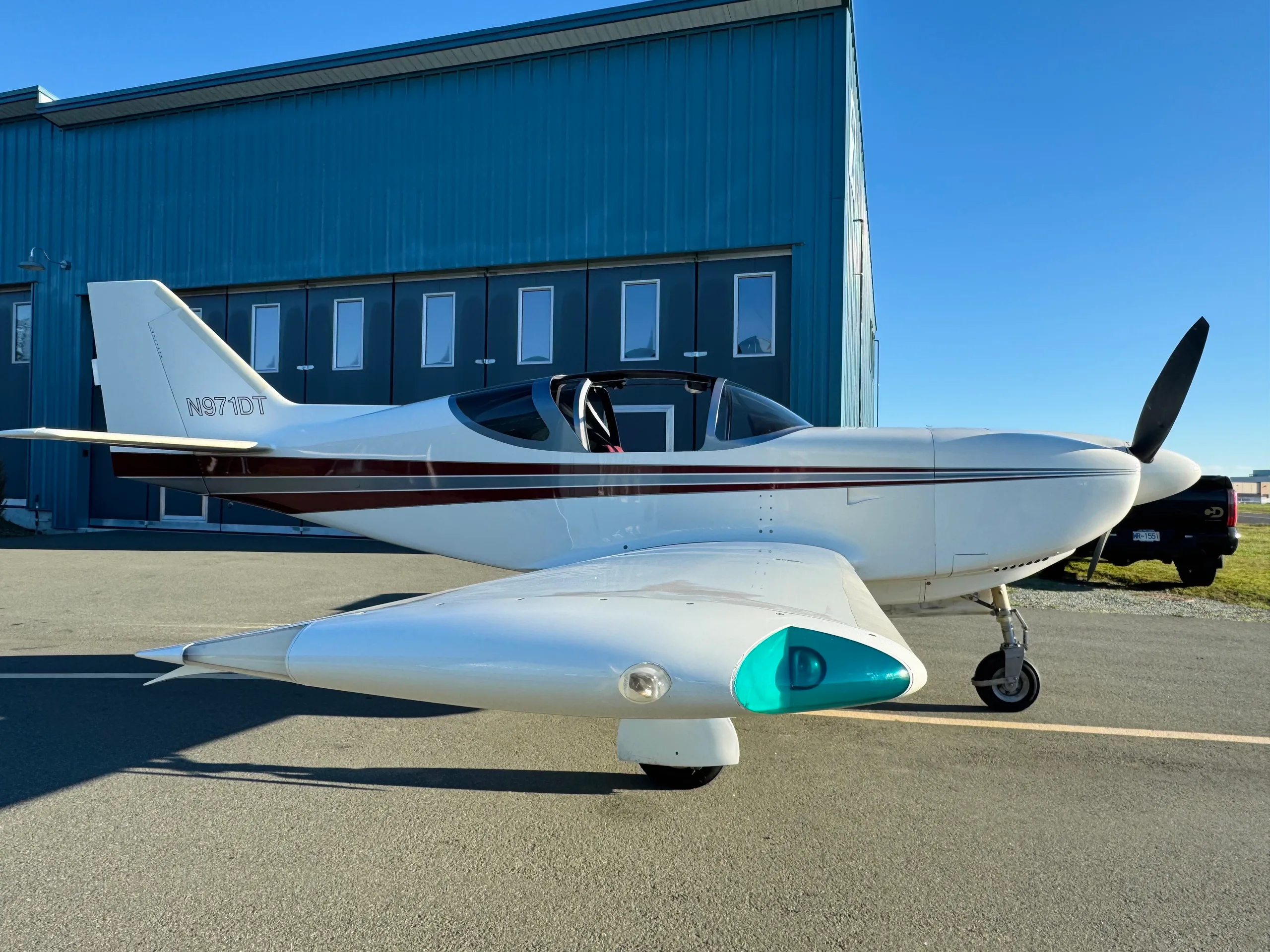 A side view of a white single-engine aircraft with brown and black stripes, registration N971DT, parked on an tarmac in front of a blue hangar.