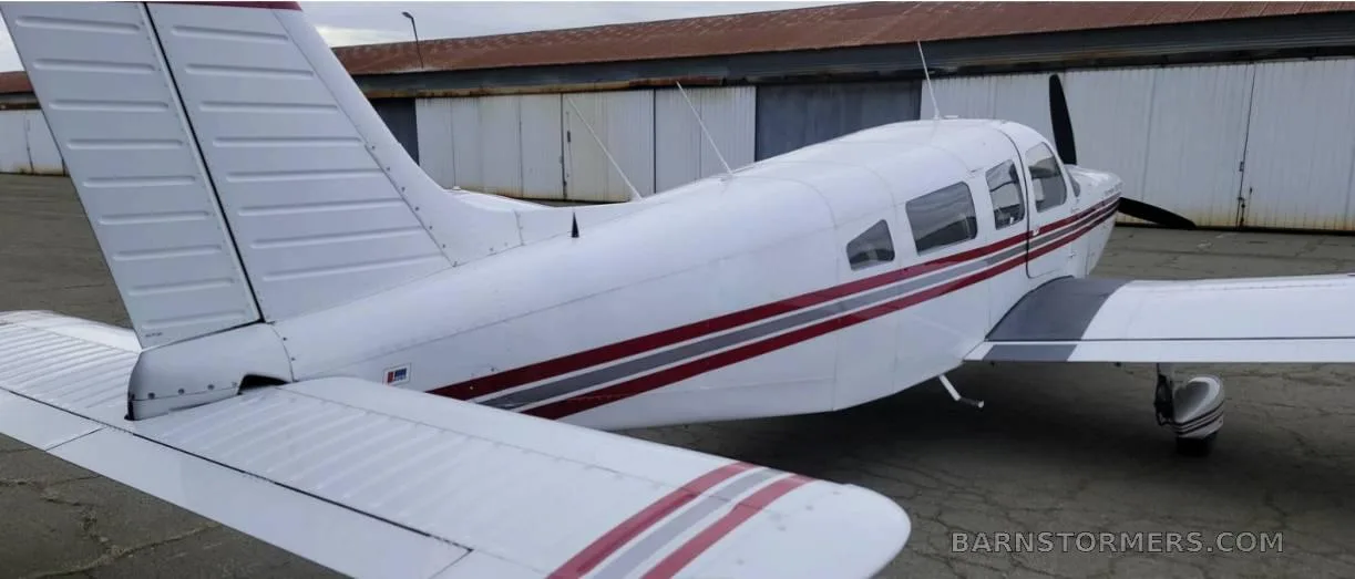 A white Piper Cherokee Six 300 aircraft with red and grey stripes, seen from the rear-right side, parked on an asphalt apron next to a hangar.