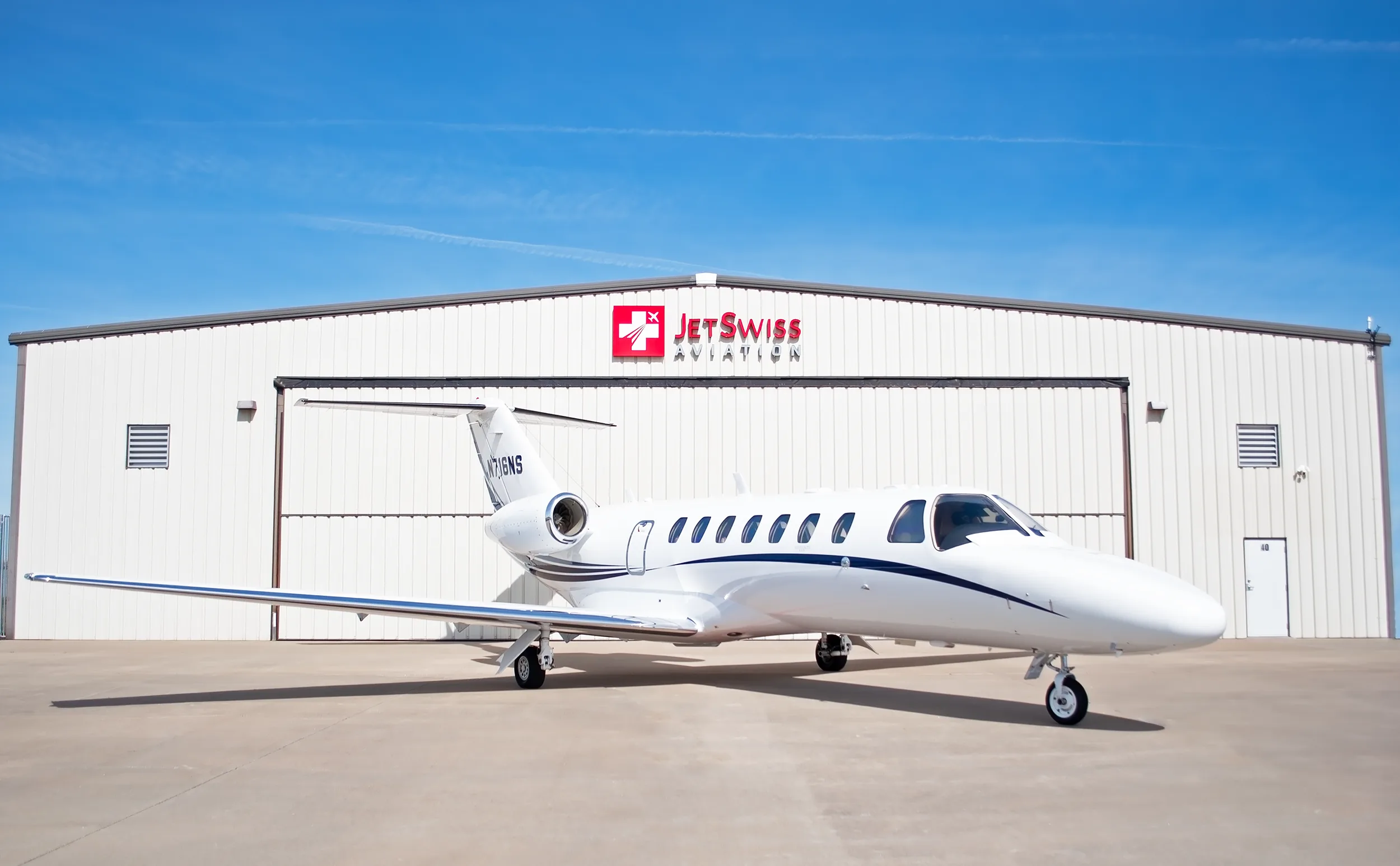 A white Cessna Citation business jet with blue stripes, registration N716NS, parked on an tarmac in front of a hangar under a clear blue sky.