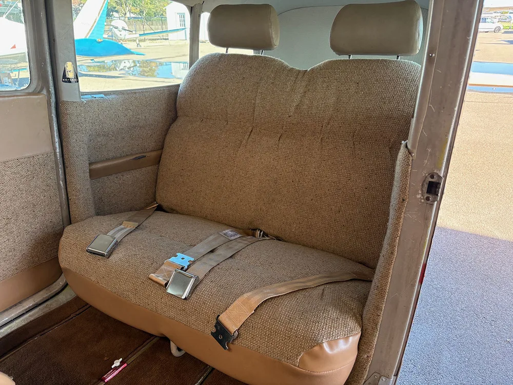 The rear bench seat of an aircraft cabin, upholstered in brown fabric and tan vinyl, with seatbelts visible.
