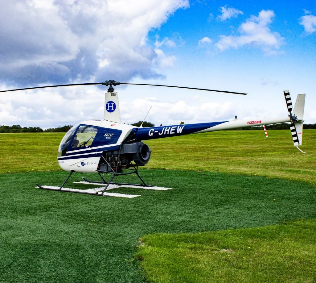 A white and blue Robinson R22 Beta helicopter (G-JHEW) with "Heli Air" branding parked on a grass helipad.