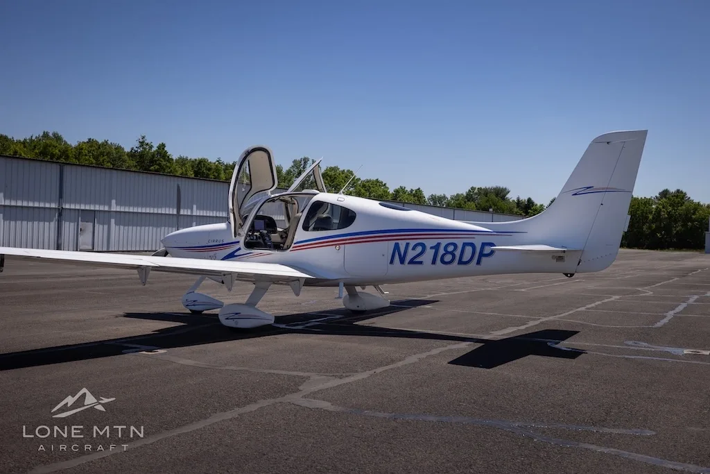 Rear-left view of a white Cirrus aircraft (N218DP) with blue and red stripes, doors open, parked on an airfield.