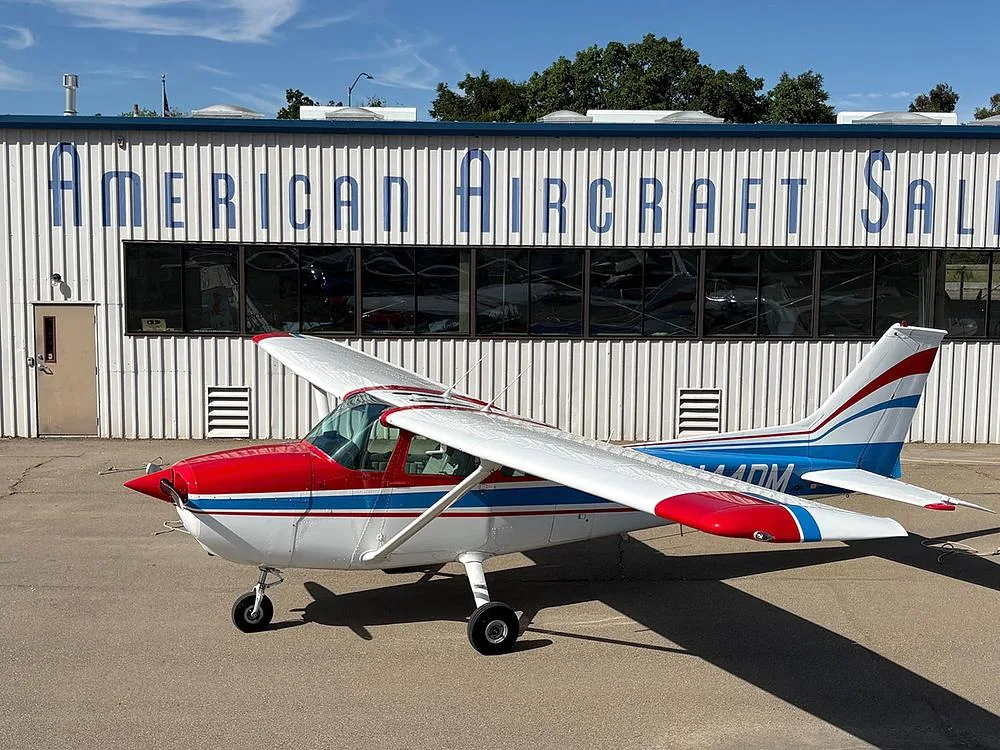 A white Cessna 172 with red and blue stripes, registration N44DM, parked in front of a building labeled "AMERICAN AIRCRAFT SALES".
