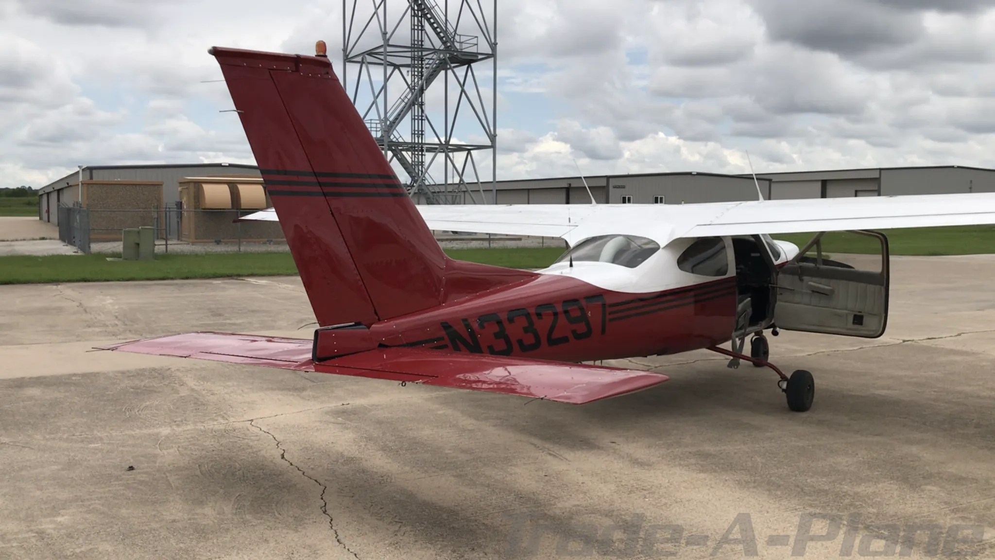 A rear-side view of the red and white N33297 aircraft with its door open, showing the tail registration, parked on an airfield with a metal tower in the background.