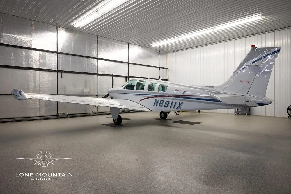 A white Beechcraft Bonanza A36 aircraft with blue and red stripes, viewed from the side in a hangar.
