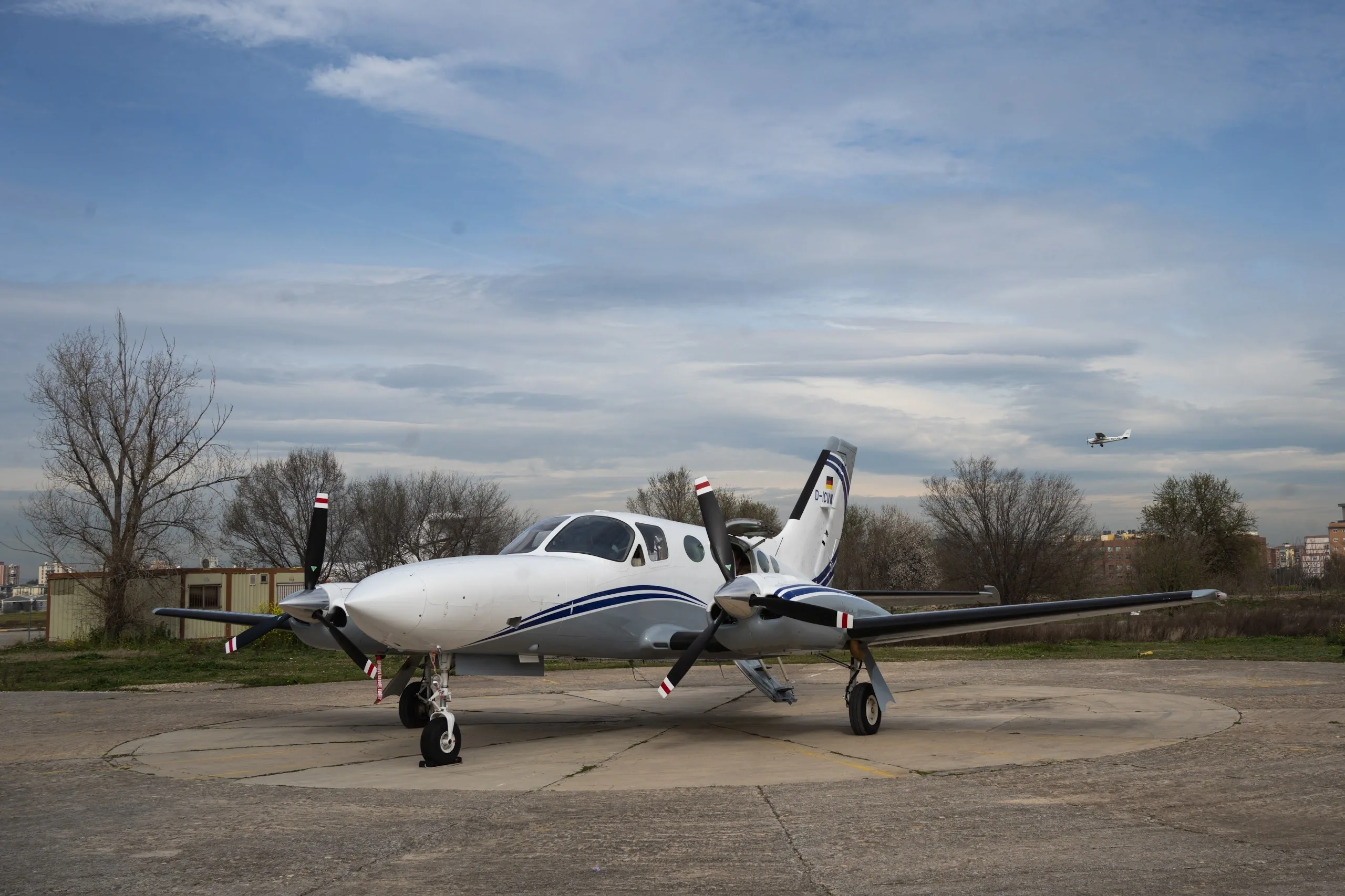 A white and grey twin-engine propeller aircraft with blue stripes parked on a concrete pad, with another small aircraft flying in the distance.