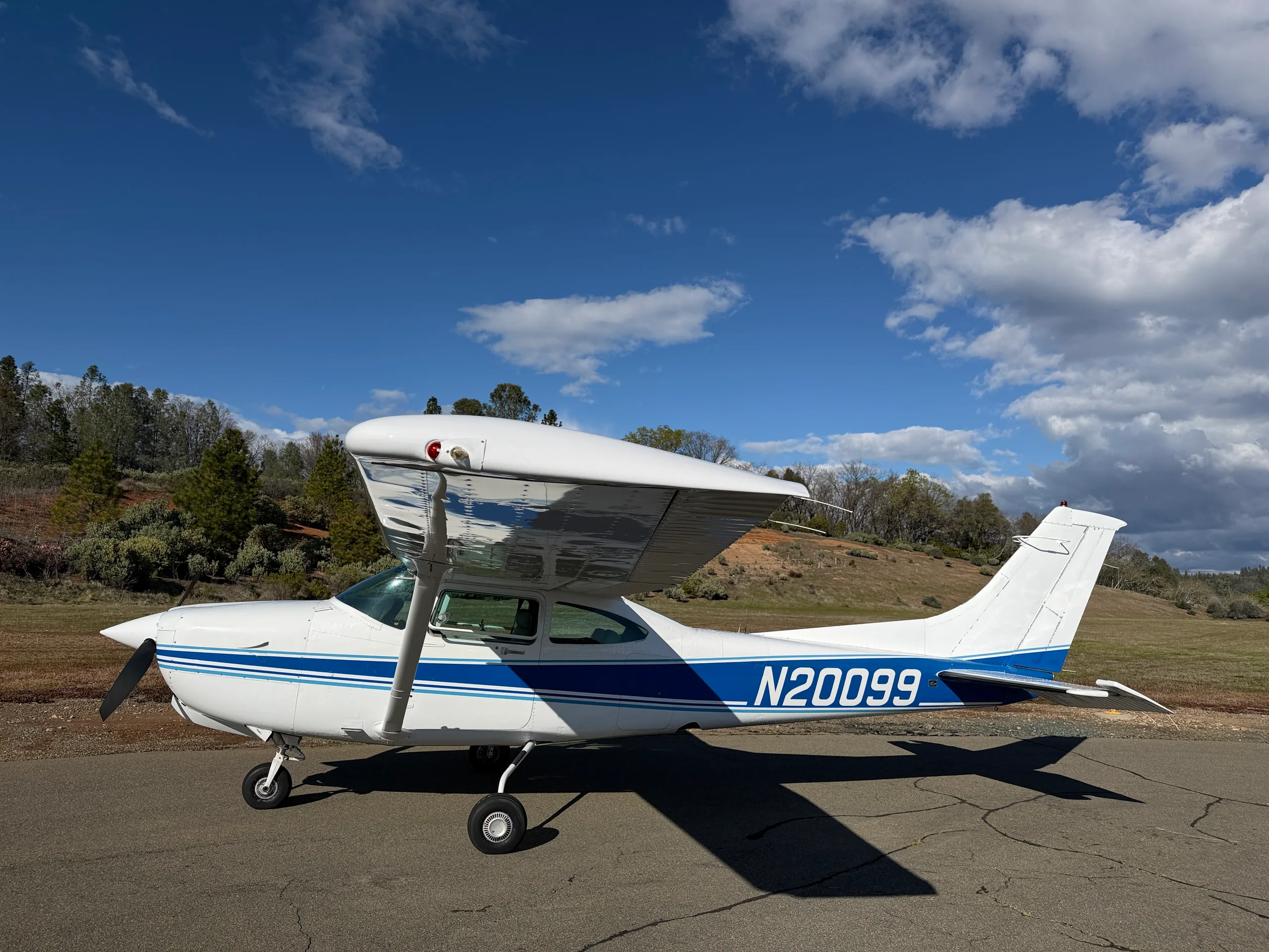 A white single-engine propeller aircraft with blue stripes, registration N20099, parked on asphalt with a hilly, treed background under a partly cloudy sky.