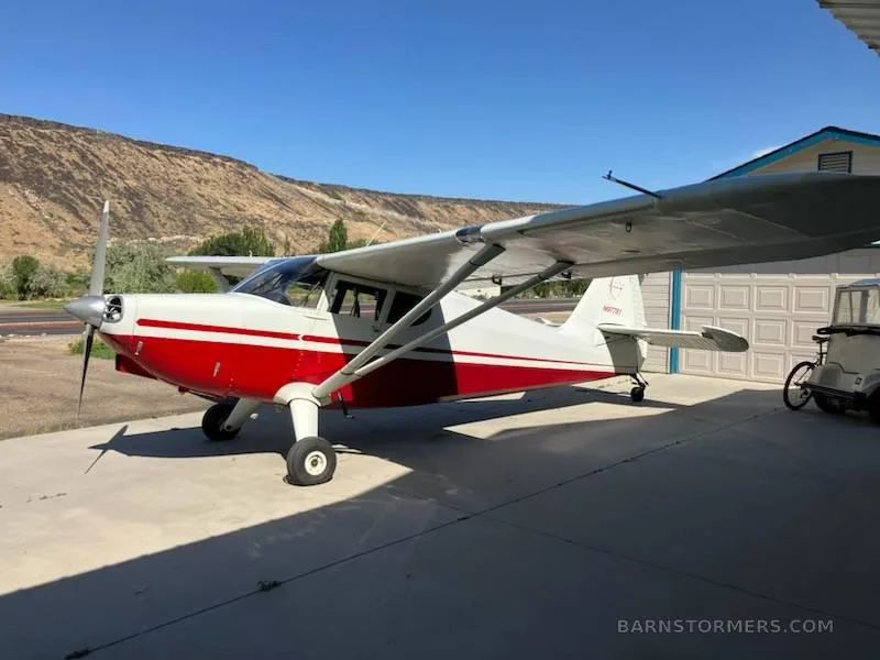 A red and white high-wing propeller aircraft parked on a concrete pad with mountains in the background.